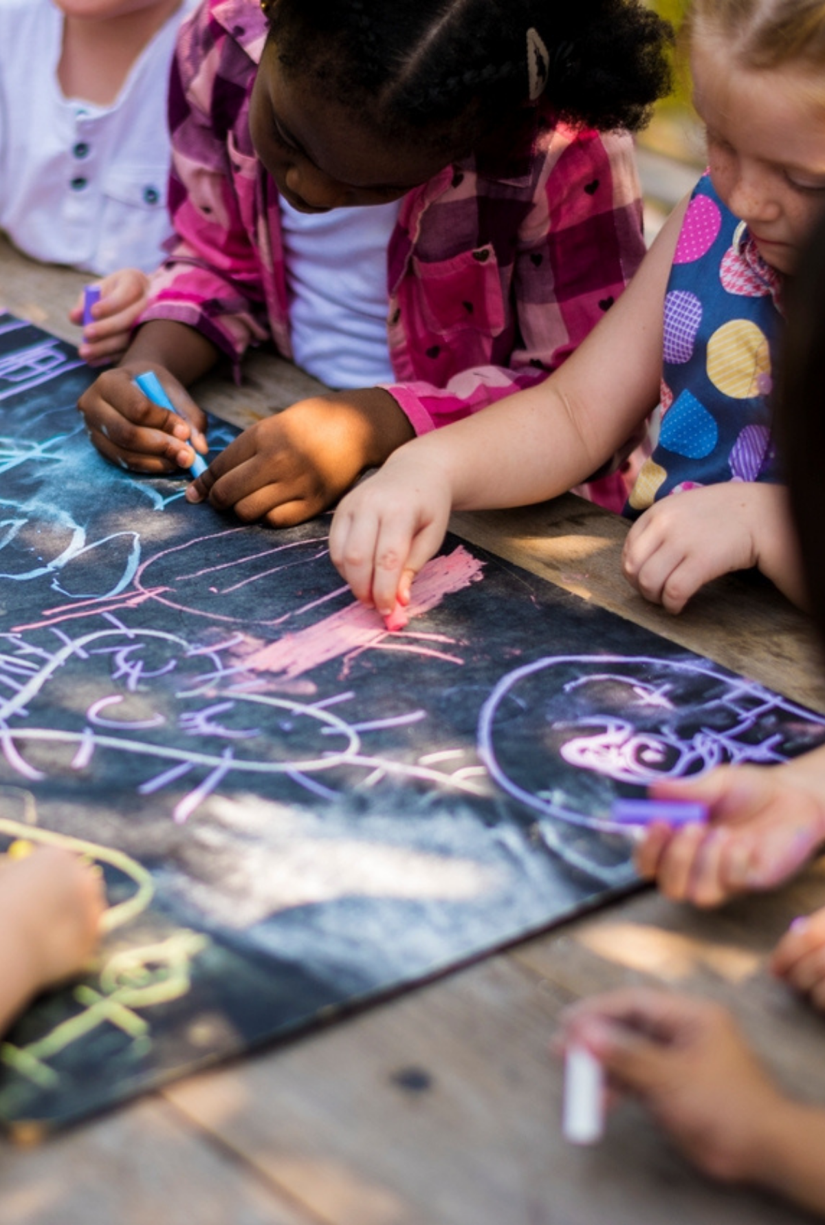 A group of children gathered around a large chalkboard outdoors, drawing bright shapes and figures together in a collaborative art activity.