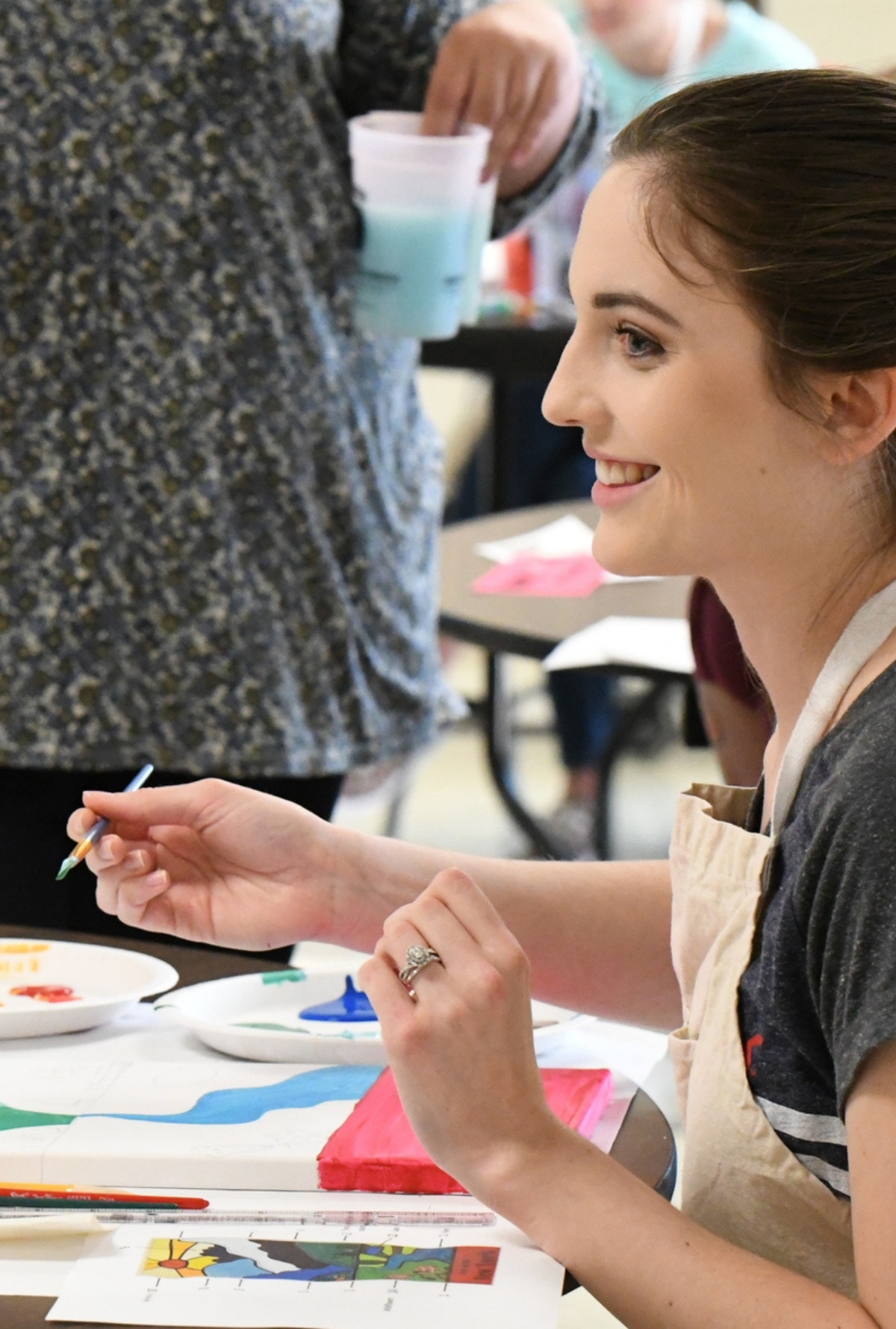 A young woman seated at a table during a community art workshop, smiling as she paints, surrounded by brushes, palettes, and other participants.